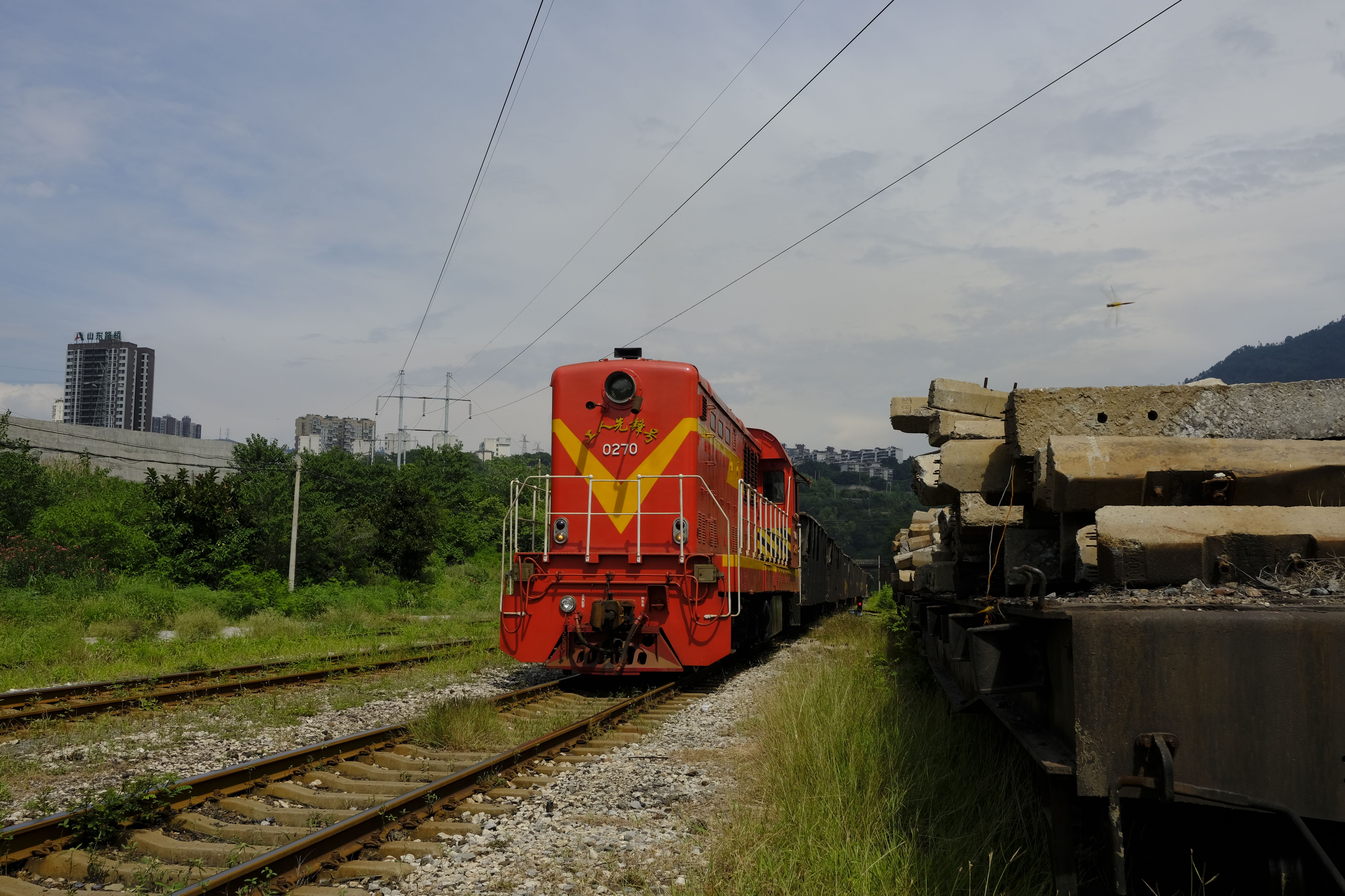 A vintage red train travels on a railway track under an overcast sky through a town with industrial elements.