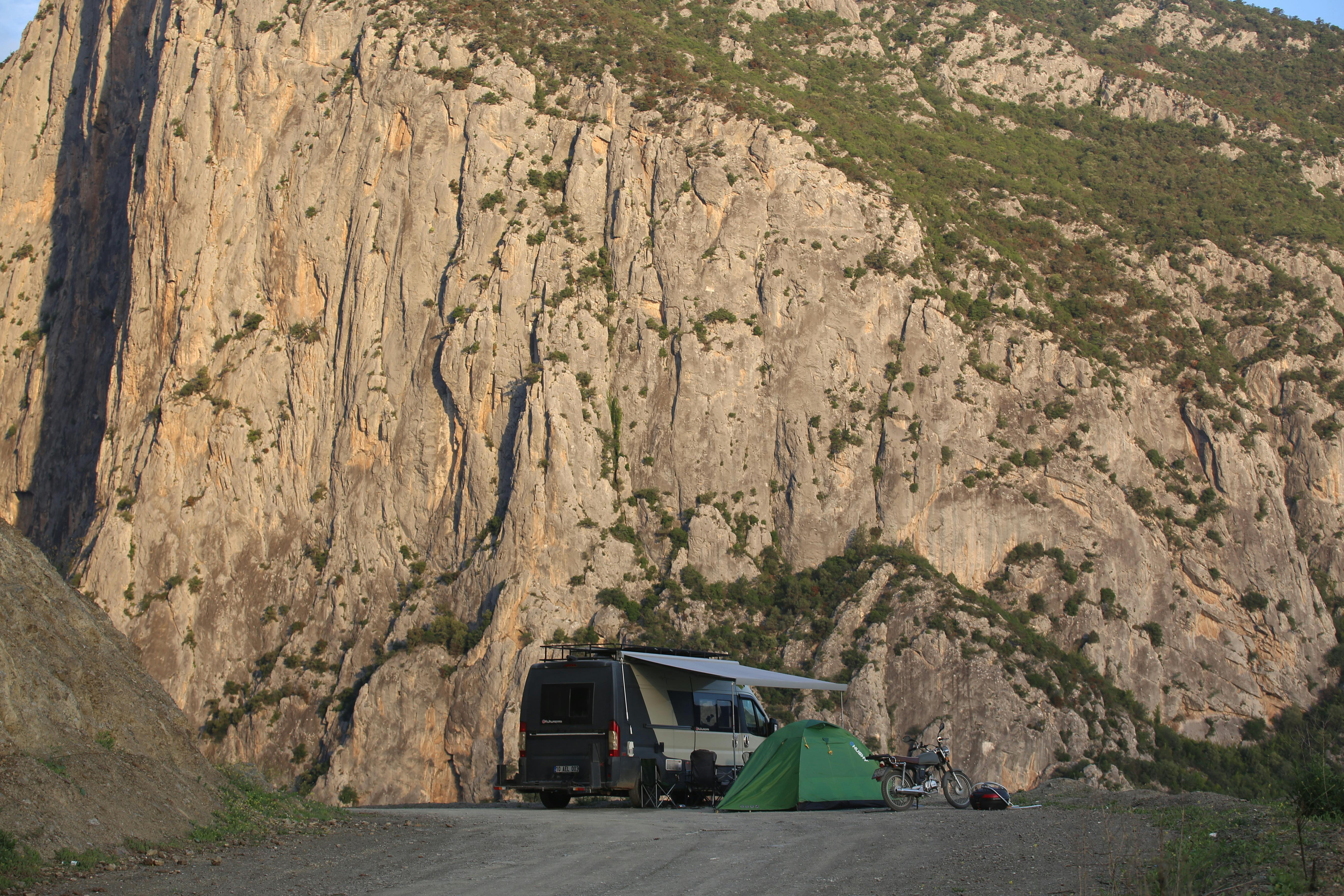 Van and tent set up for camping beside towering cliffs on a sunlit day.