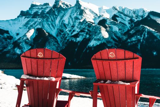 Scenic winter view of red chairs facing snowy mountains by a lake in Banff National Park, Canada.