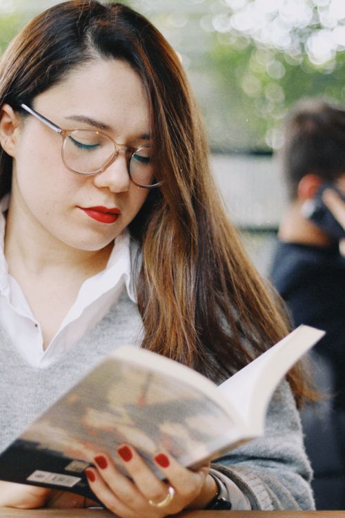 Free Woman with glasses and long hair reading a book in a relaxed cafe setting. Stock Photo