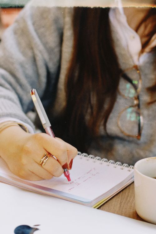 Free Close-up of a woman's hand writing notes in a notebook with a cup of coffee nearby. Stock Photo