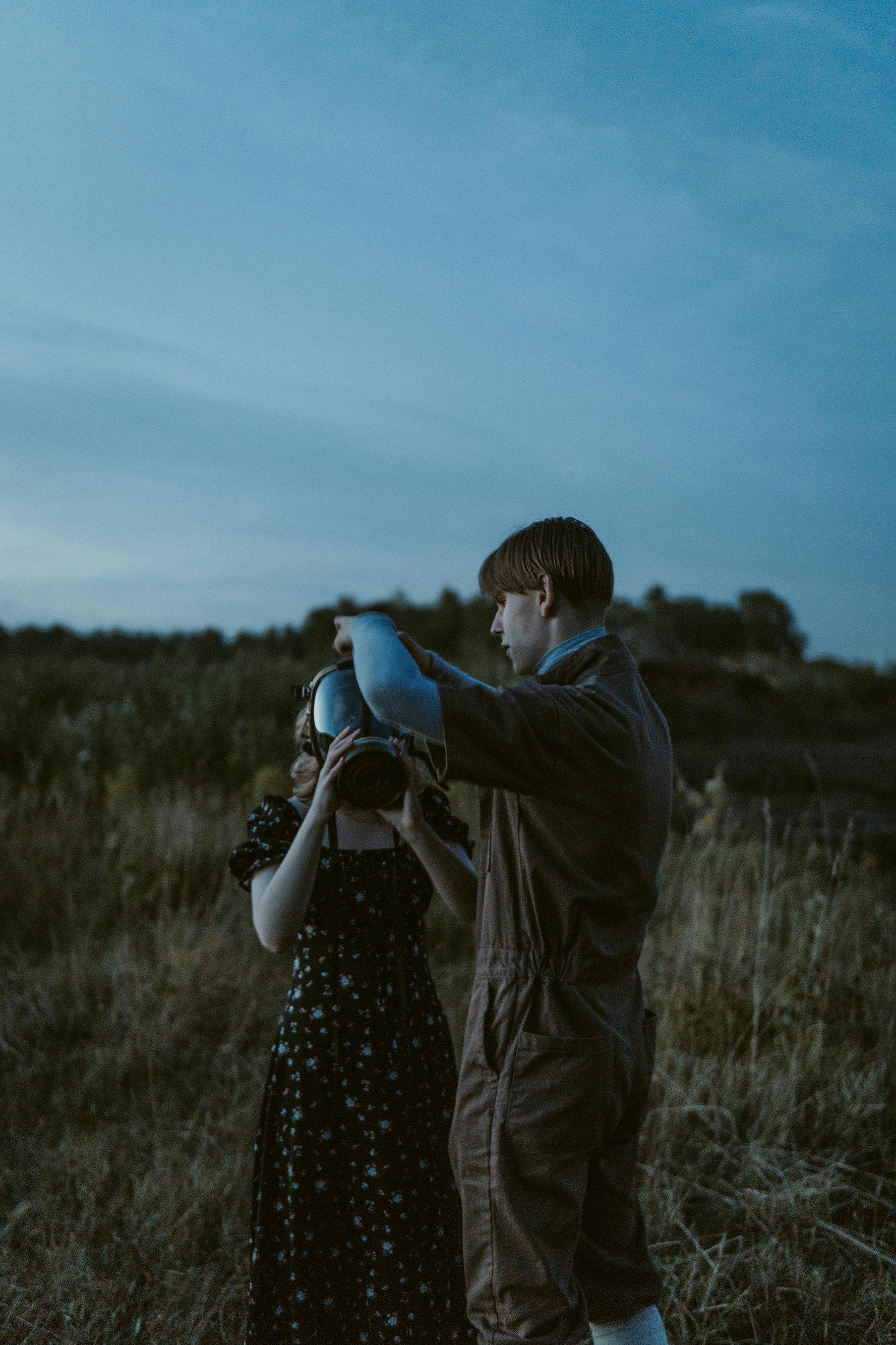 A young couple in a field at dusk with a gas mask, showcasing a conceptual theme.