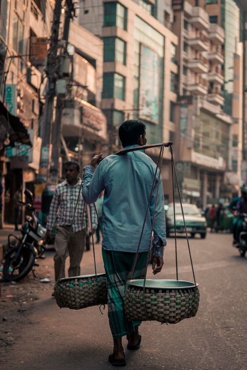 Free A man carrying traditional baskets walks down a bustling urban street, showcasing everyday life. Stock Photo