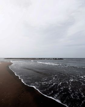 A peaceful beach scene with dark sand and a calm sea under an overcast sky.