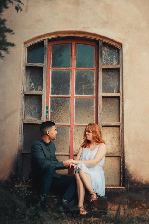 Free A loving couple sitting and smiling while holding hands in front of a rustic building exterior. Stock Photo