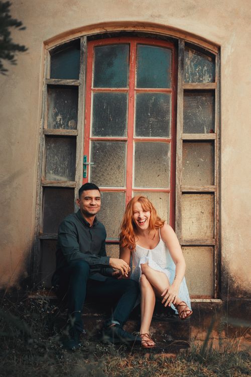 Free Smiling couple sitting in front of a rustic windowed wall, exuding joy and warmth. Stock Photo