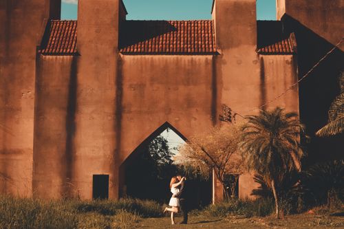 Free A couple embracing in front of a sunlit ancient wall surrounded by green foliage, evoking romance and warmth. Stock Photo