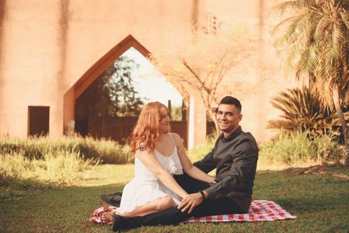 Free A couple enjoying a sunny day with a picnic on the grass, surrounded by lush greenery and architecture. Stock Photo