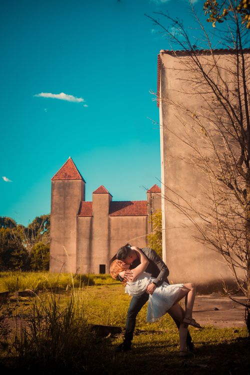 Free A couple shares a romantic kiss outdoors near a medieval-style building under a clear blue sky. Stock Photo