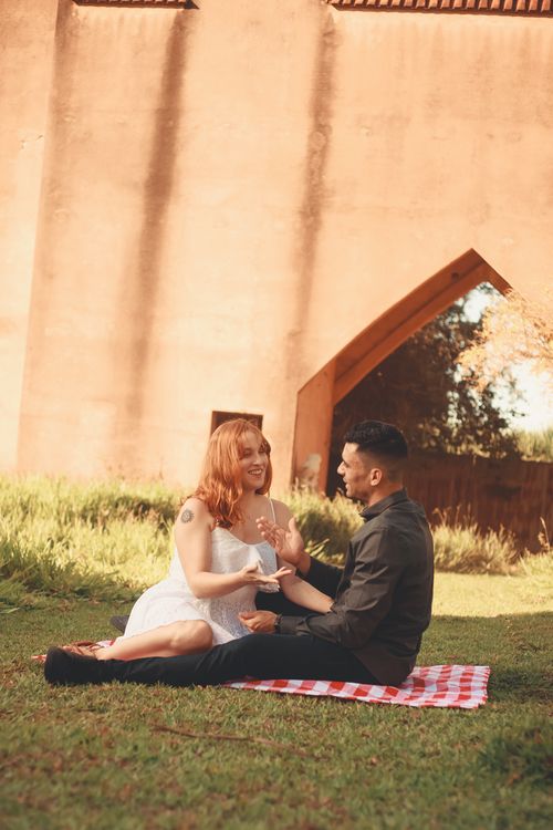Free Couple enjoying a picnic on a sunny day near a historical archway, sharing smiles and conversation. Stock Photo
