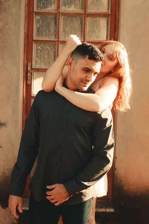 Free Young couple standing together, embracing outdoors by a glass-door, in warm light. Stock Photo