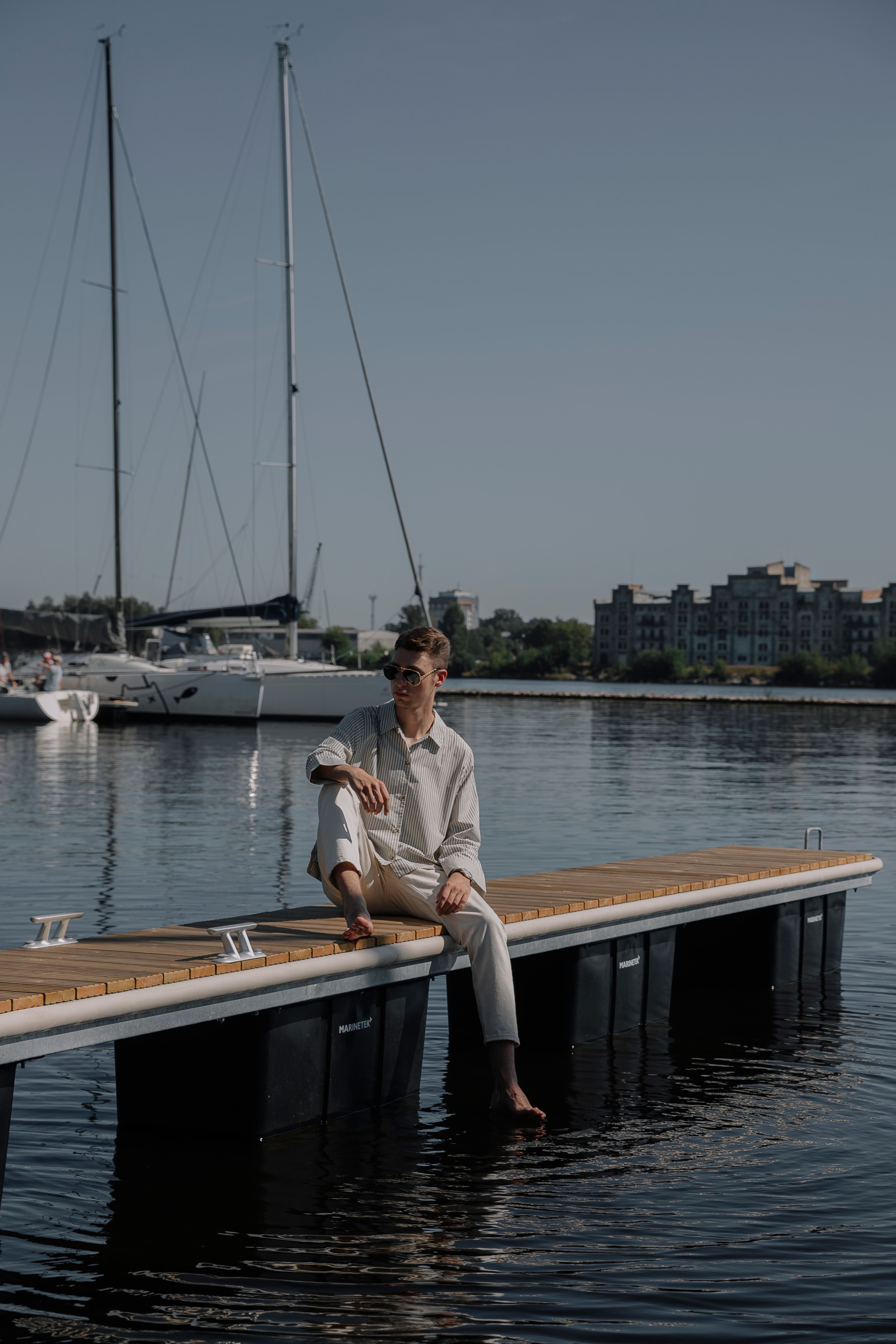 A fashionable man in white clothes sitting on a pier surrounded by yachts.