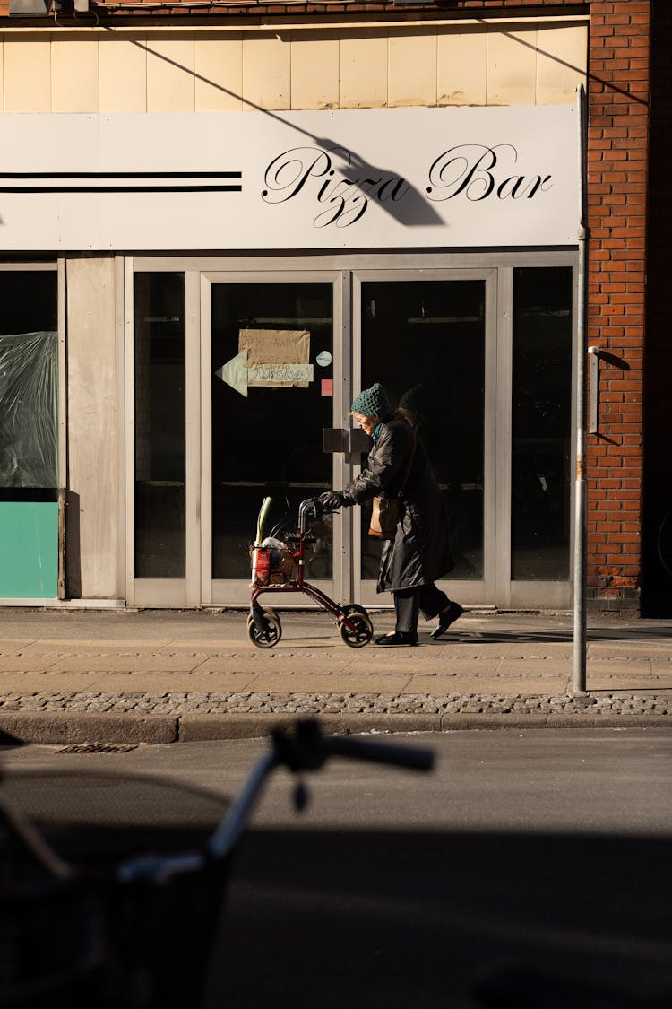 Candid Photo Of An Elderly Woman Walking On A Sidewalk Next To A Pizza Bar 