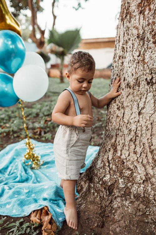 Free A young boy standing barefoot next to a tree with balloons and a blanket outdoors. Stock Photo
