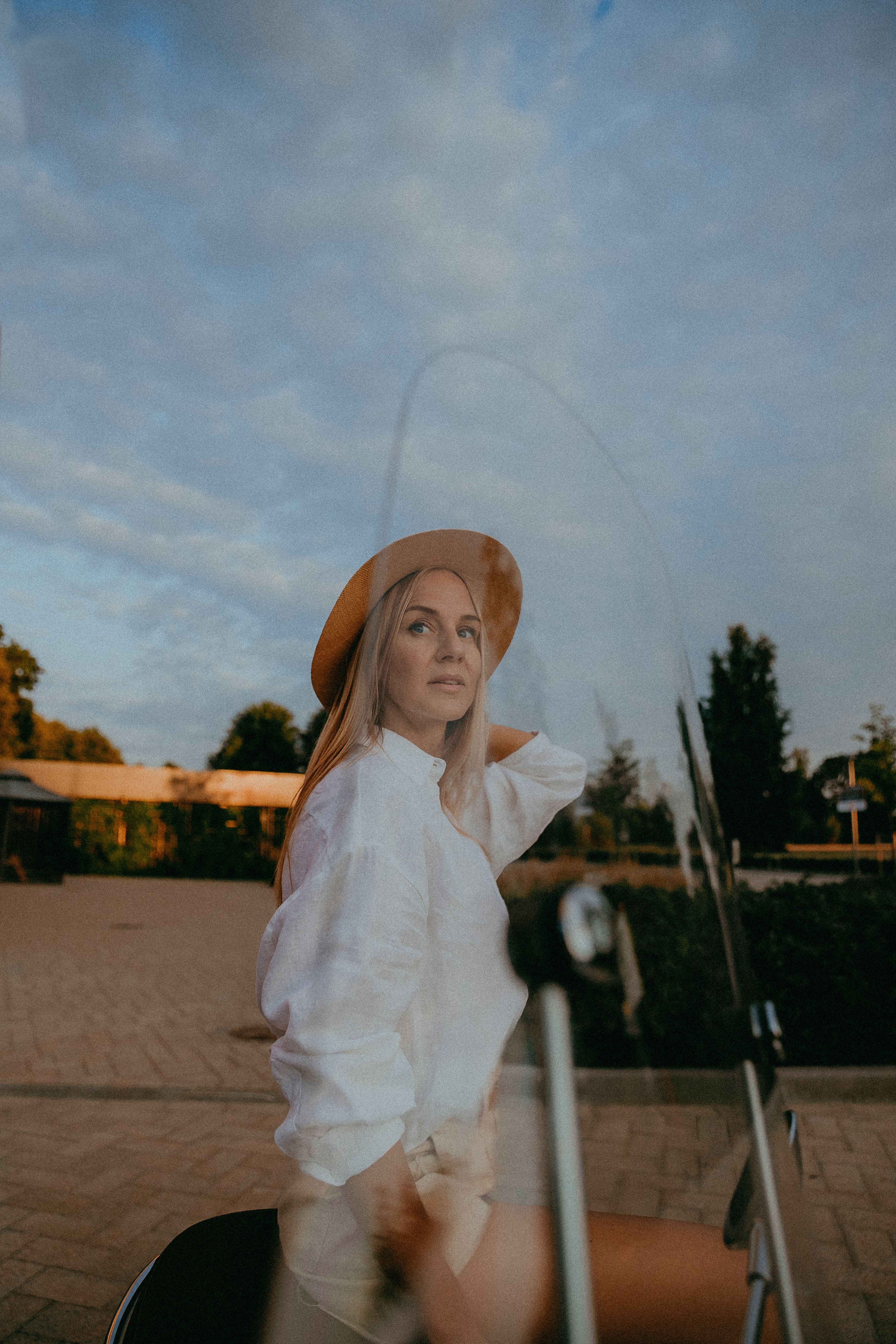 A fashionable woman in a white shirt and hat poses by a scooter at sunset.