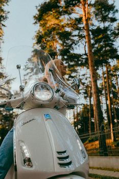 Blonde woman riding a white Vespa scooter in a sunny forest setting, enjoying leisure time outdoors.