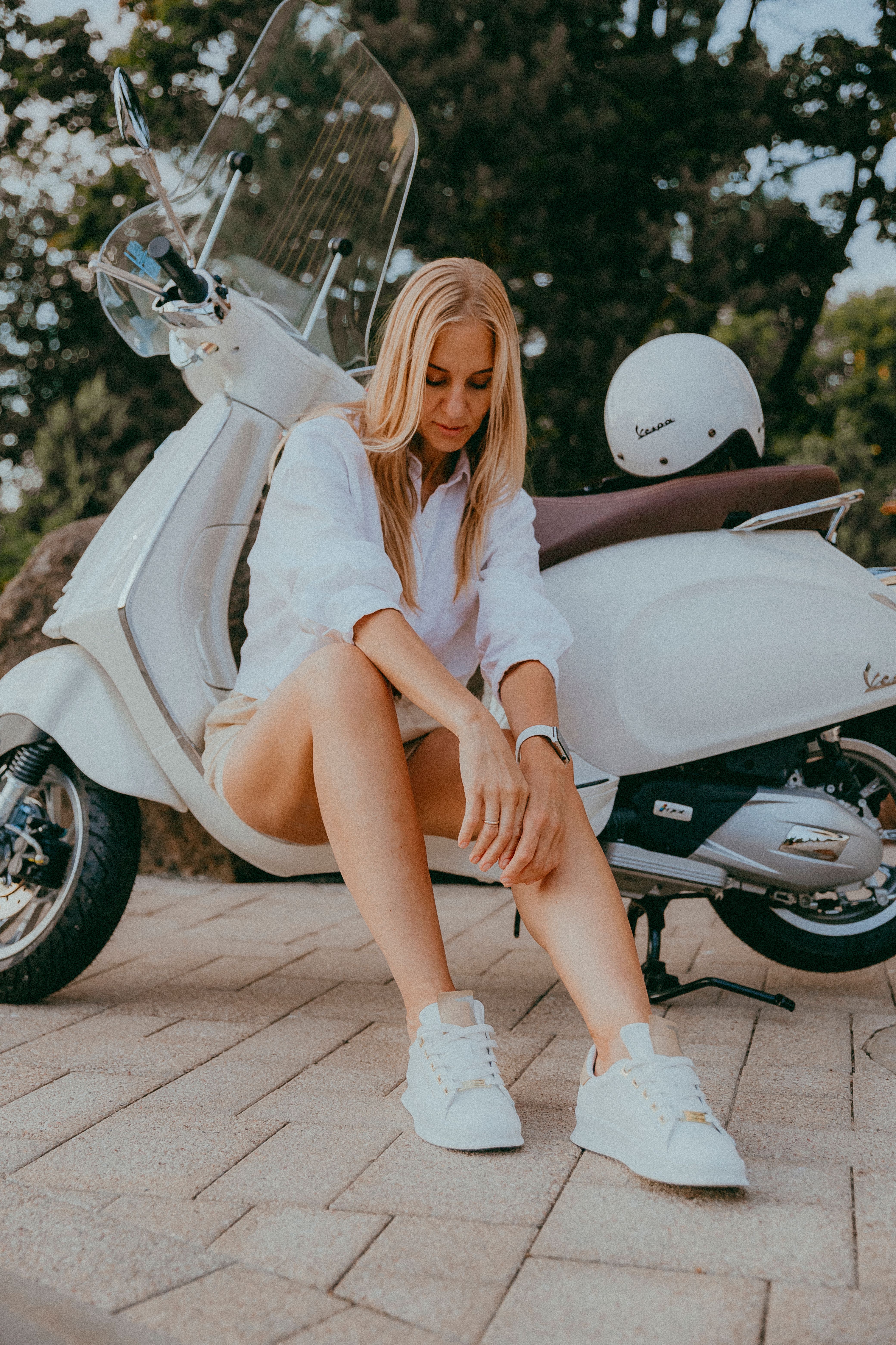 Blonde woman in white shirt sitting by a Vespa scooter outdoors.