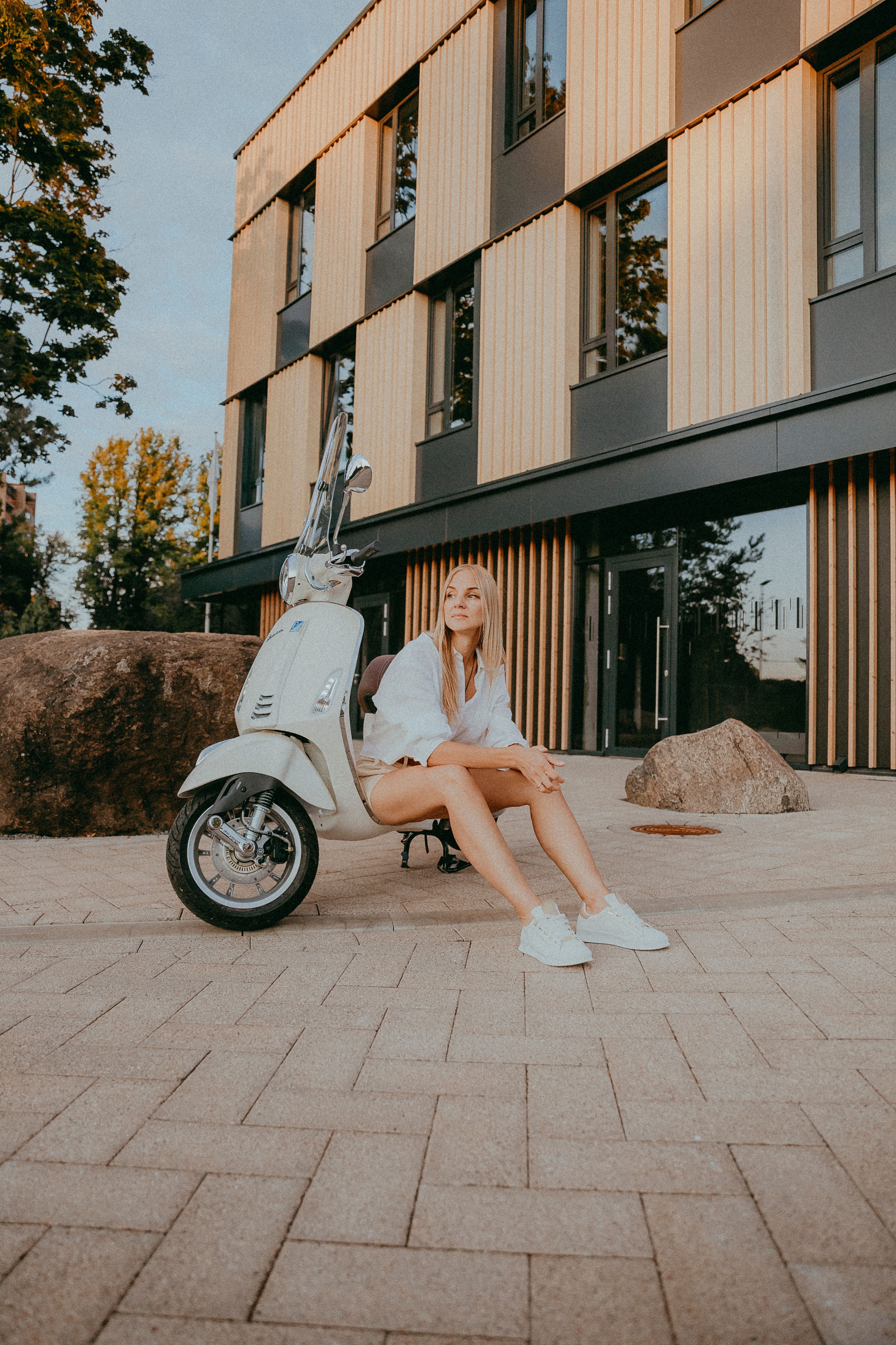 Stylish woman sitting beside a scooter in an urban setting, showcasing modern fashion.