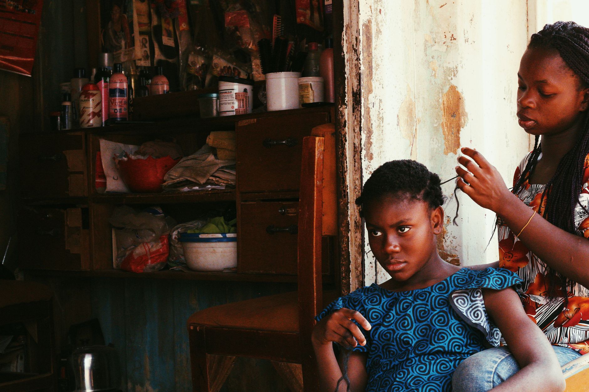 A woman braiding a girl's hair at home, sections held in her hands.