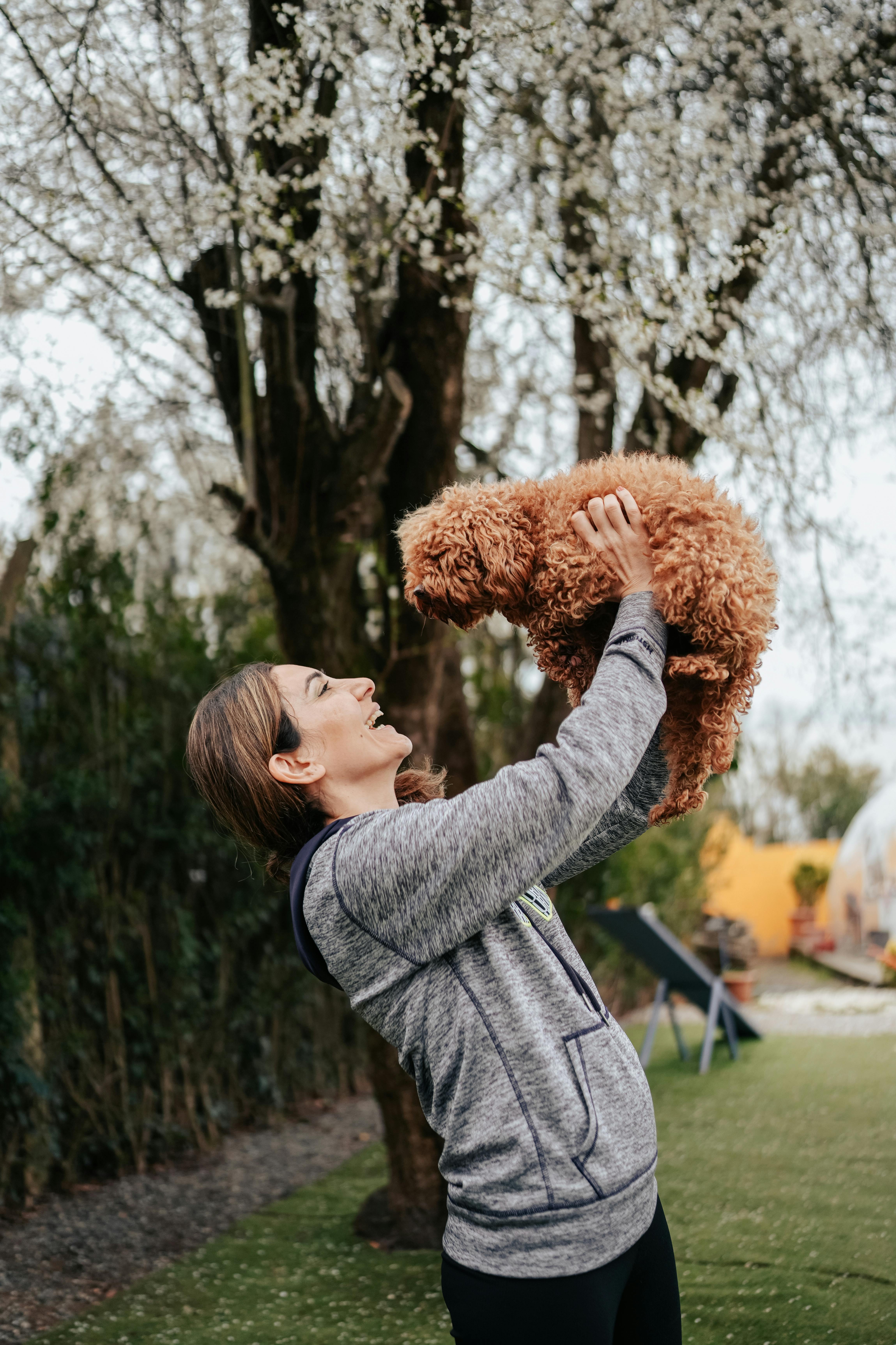 Woman happily lifts her fluffy dog amidst blooming trees in a park, capturing a joyful moment of pet companionship.