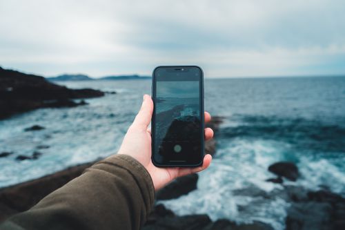 Free A hand holding a smartphone capturing waves and horizon by the seaside. Stock Photo