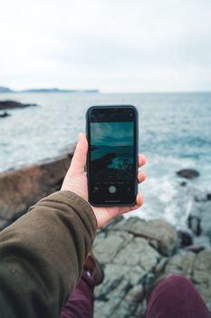 A hand holding a smartphone capturing a scenic ocean view from rocky shores.