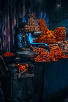 A vibrant Indian street food market stall with a vendor sitting amid stacks of traditional snacks in Canning, WB, India.
