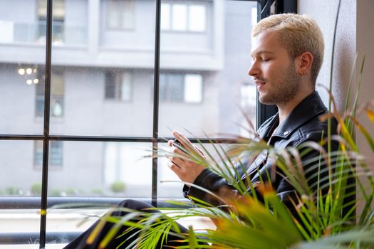 Young Man in a Leather Jacket Sitting by the Window and a Plant 