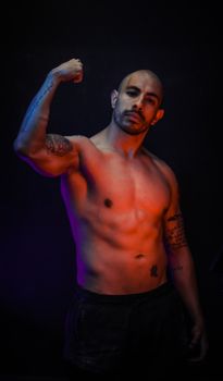 Shirtless muscular man with tattoos posing confidently in dramatic studio lighting.