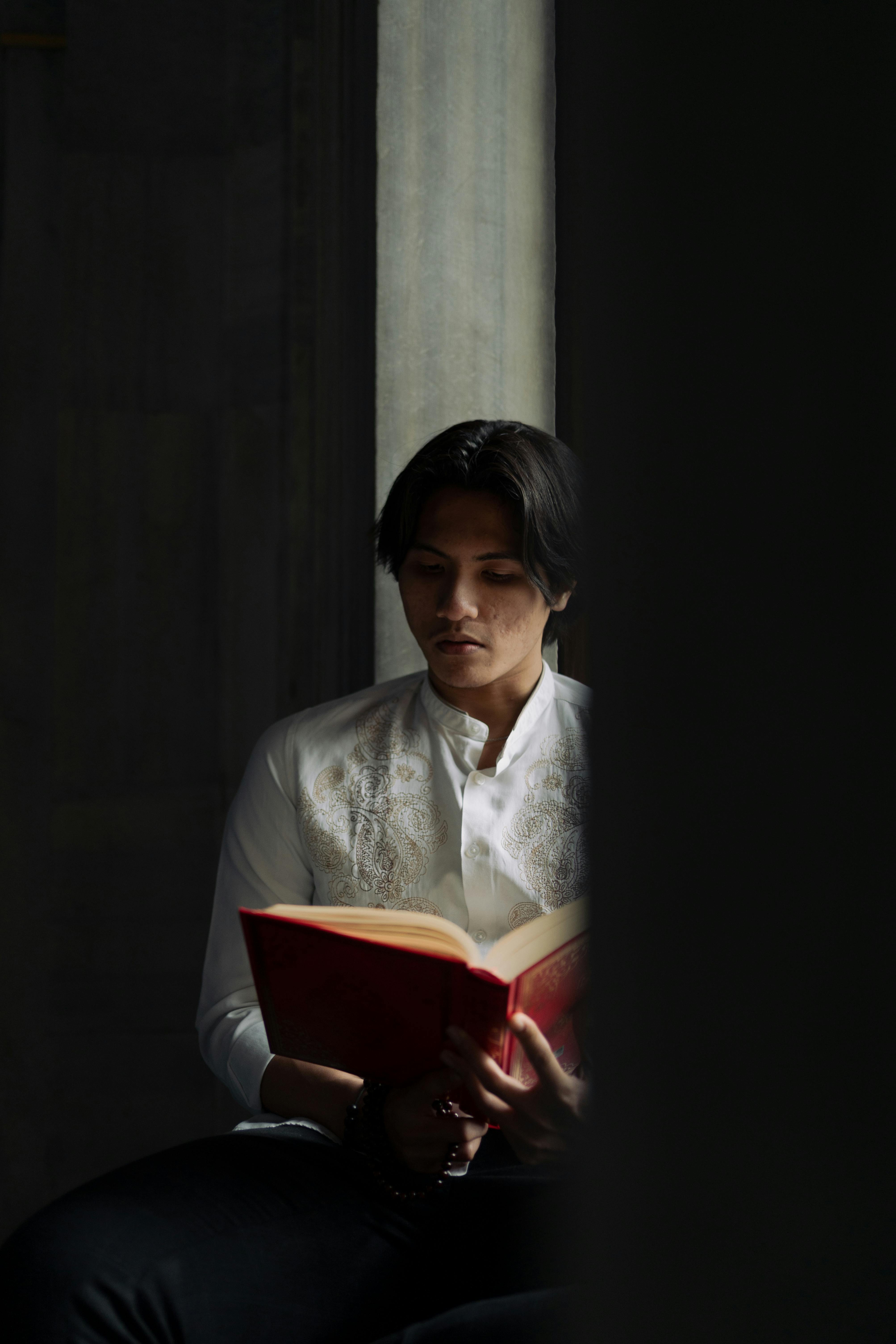 A young man in traditional attire thoughtfully reading a book indoors.