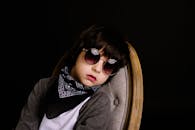 深色背景的椅子上，坐着一个戴太阳镜和围巾的时尚孩子