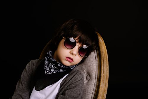 Free Portrait of a fashionable child wearing sunglasses and a scarf, sitting on a chair against a dark background. Stock Photo