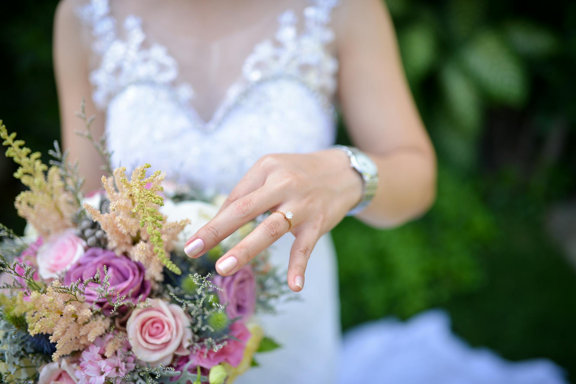 Close-up of bride's hand showcasing engagement ring with elegant wedding bouquet outdoors.