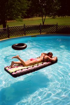 A woman in a bikini relaxes on an airbed in a sunny backyard swimming pool.