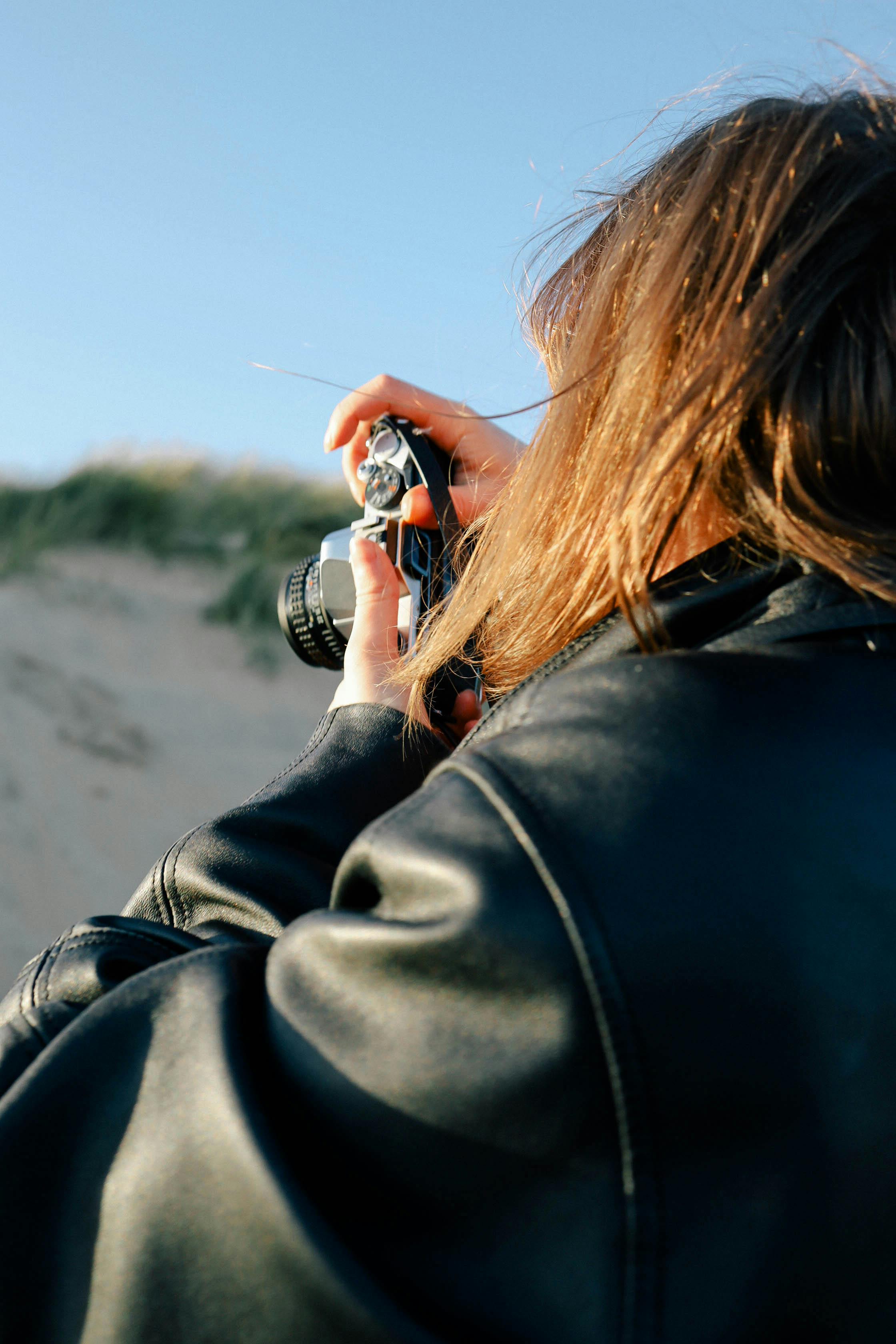 A woman with long hair holds a camera outdoors, capturing a scenic view.