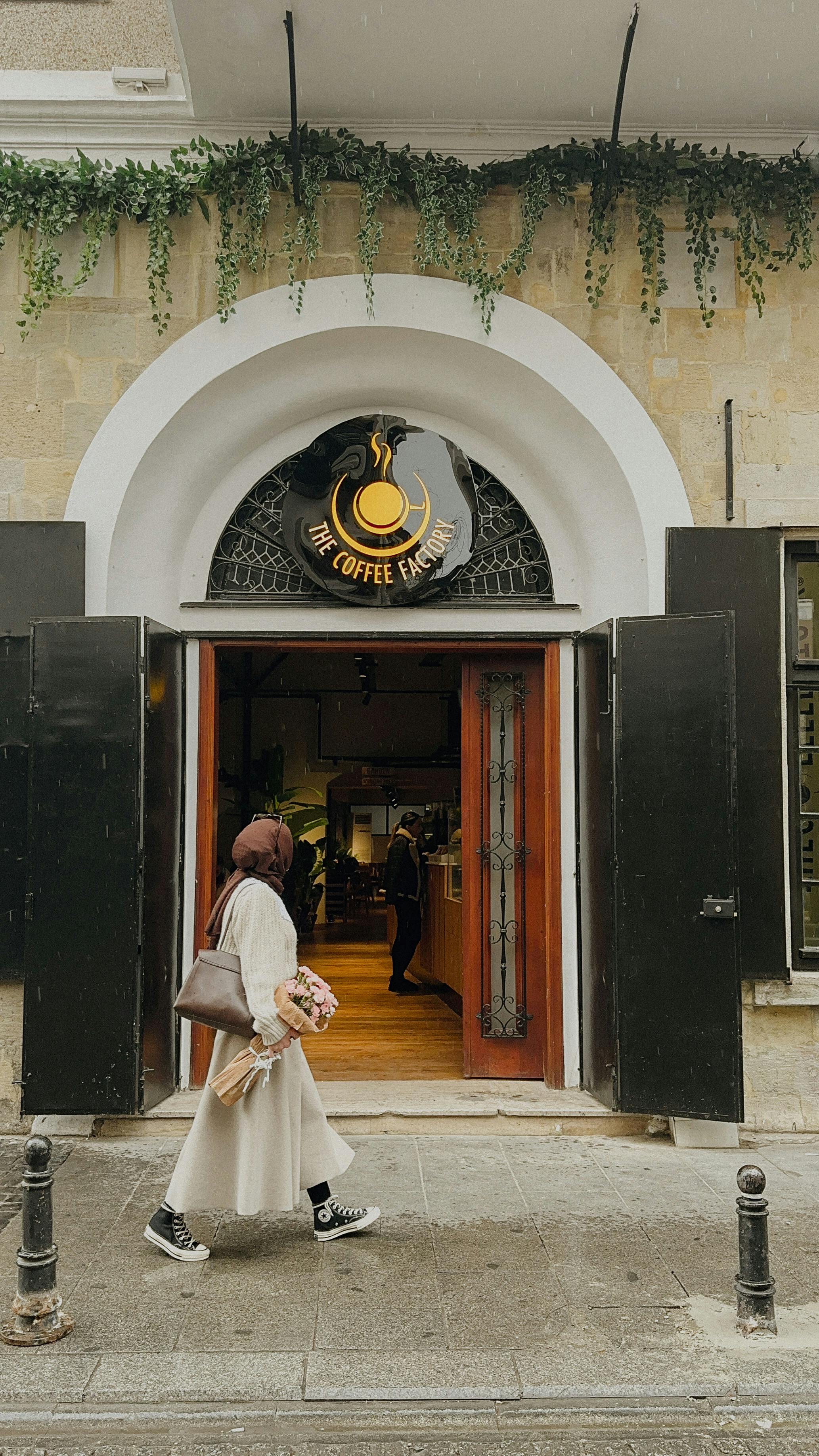 A woman in hijab walks into a café, carrying a bouquet, in an urban setting.