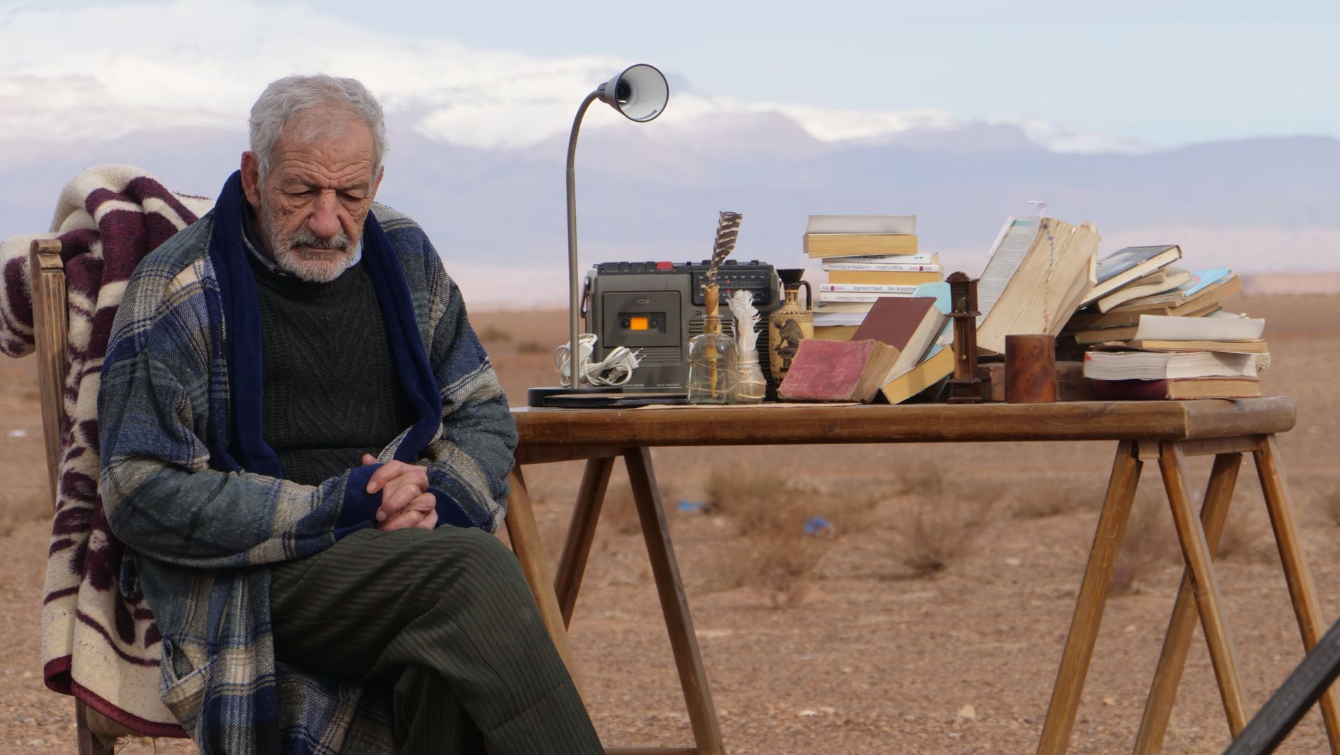 An elderly man sits beside a table filled with books and equipment in the Moroccan desert.