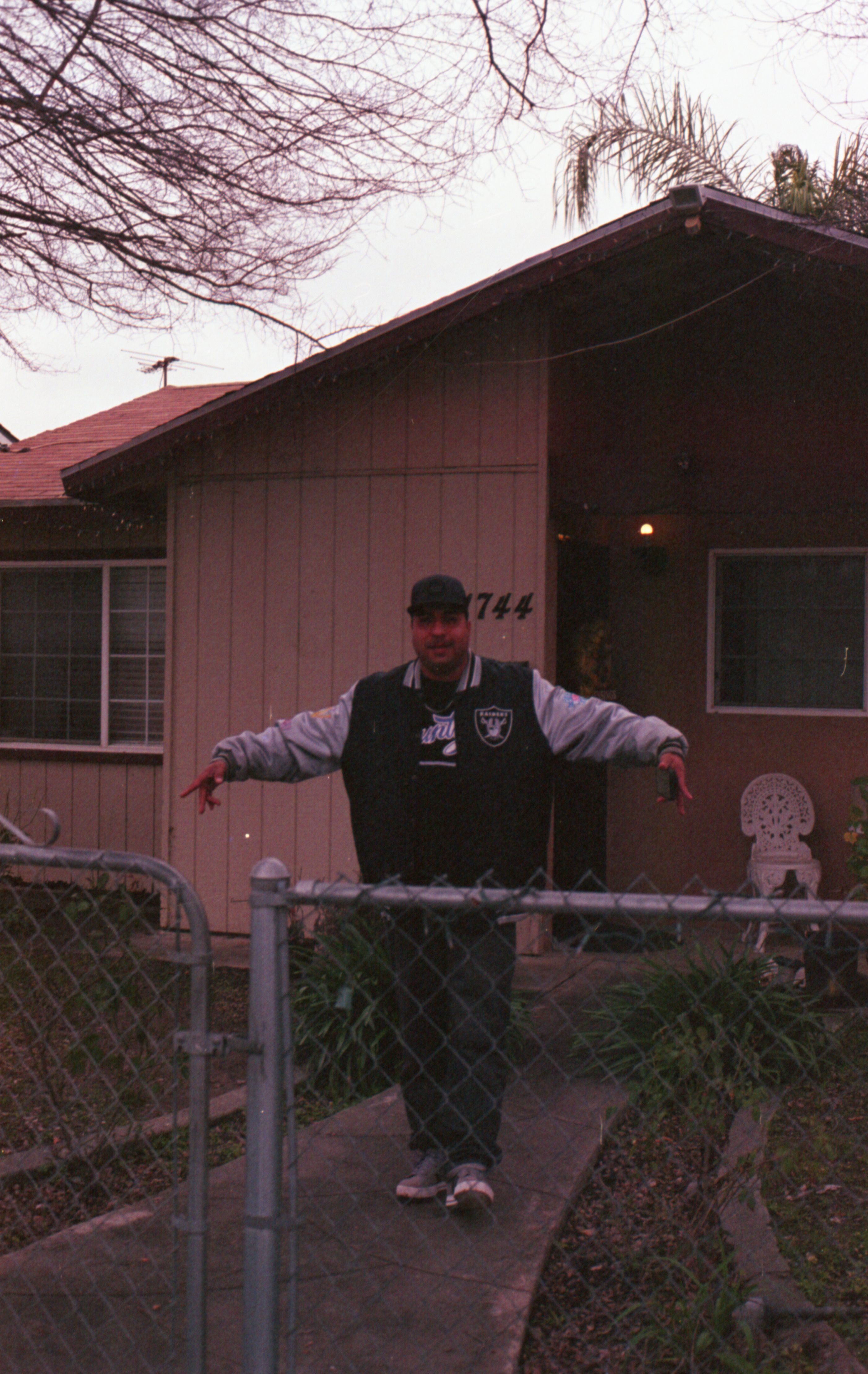A man wearing a jacket and baseball cap stands in a city yard with hands raised near a house.