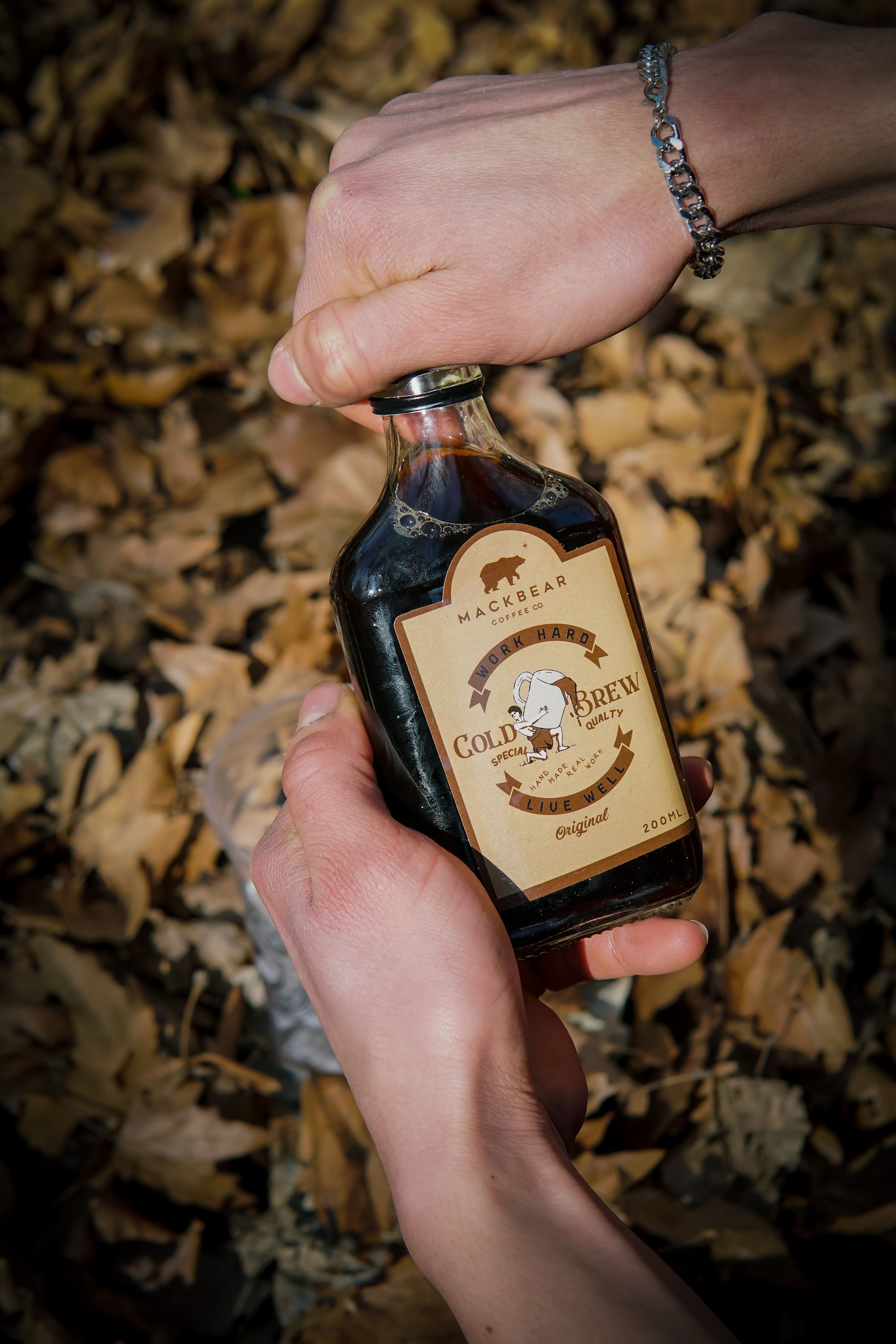 A close-up of hands opening a cold brew bottle outdoors with fallen leaves.