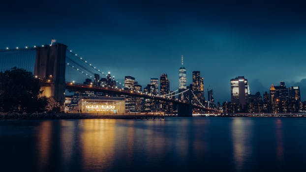 Night view of Brooklyn Bridge with illuminated New York City skyline across calm waters.