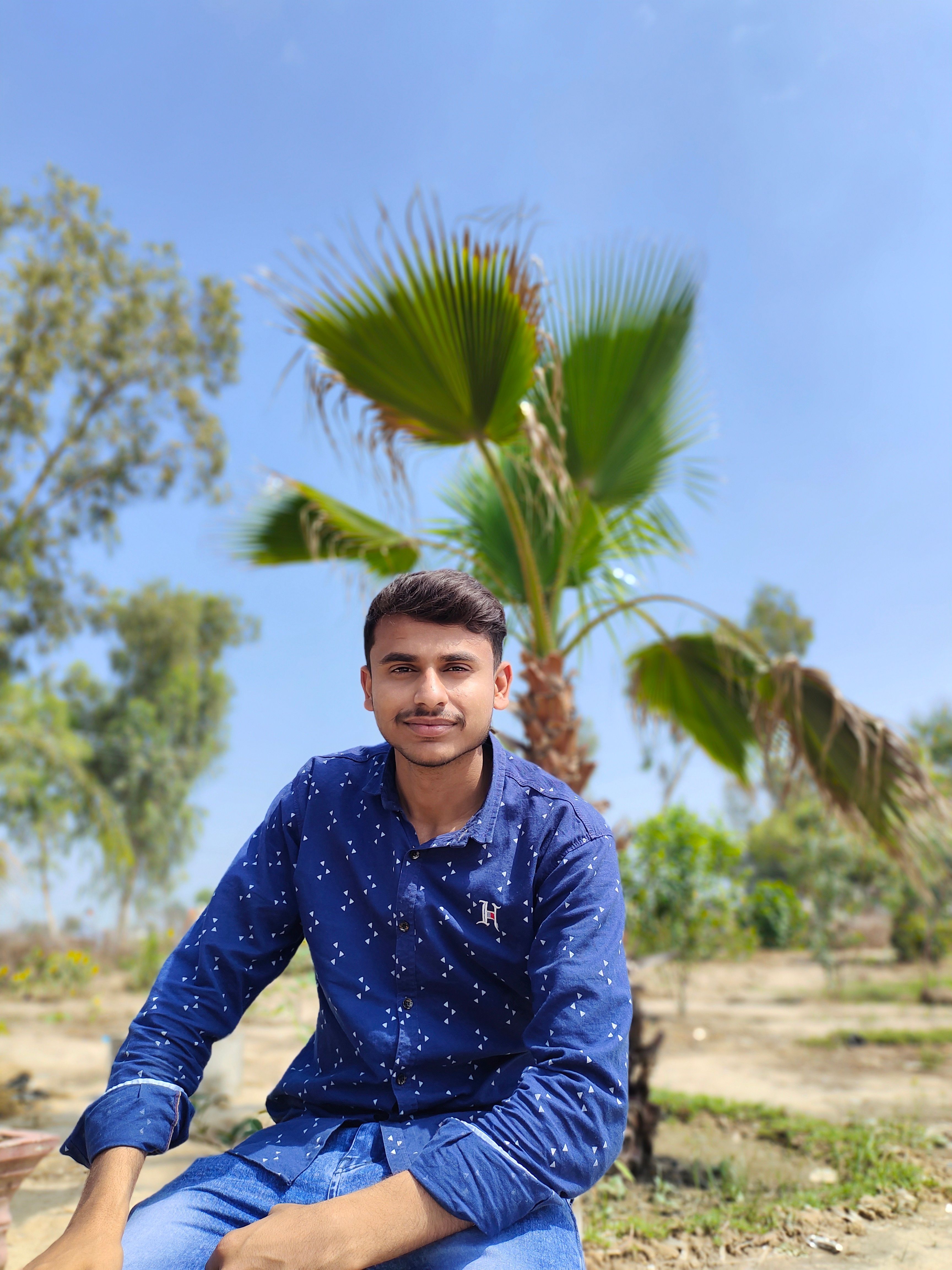 A young man in a navy shirt smiles while sitting outdoors with palm trees in Athilpur, Pakistan.