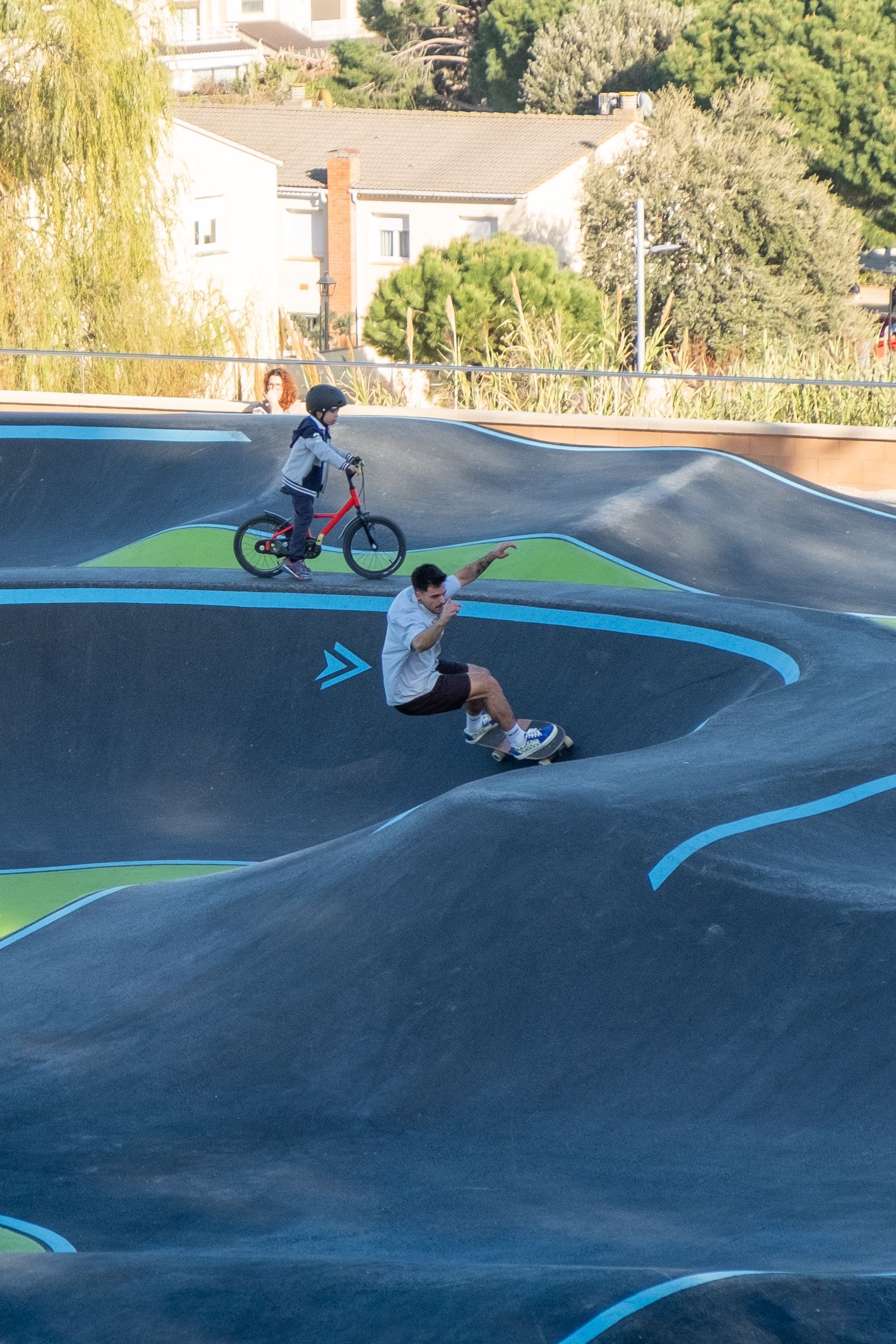A dynamic scene of a child cycling and a man skateboarding at an outdoor skatepark.