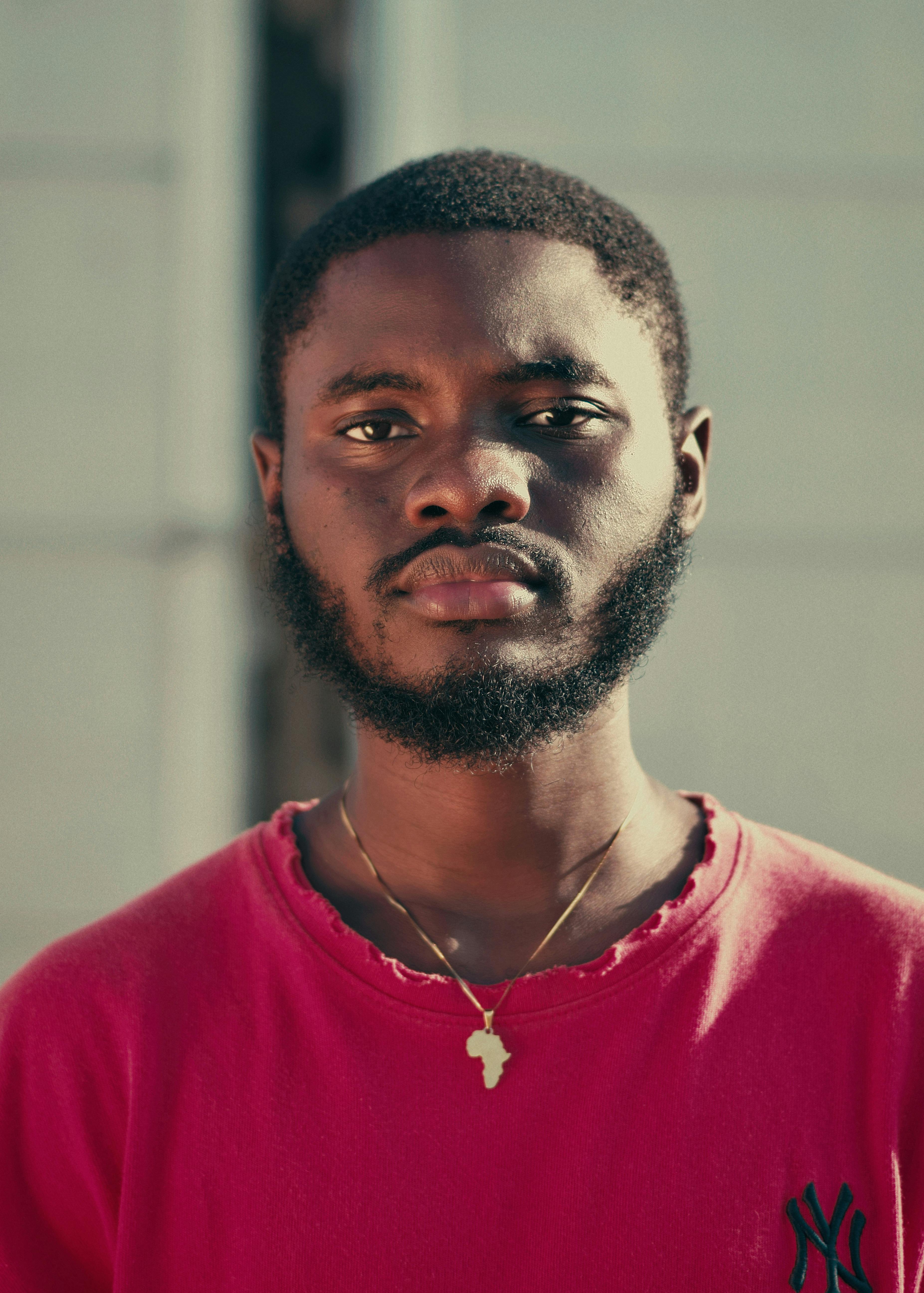 Close-up portrait of a man wearing a red shirt and necklace against a neutral background.