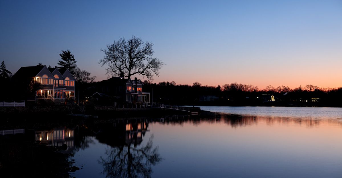 Tranquil lakeside scene at sunset, capturing reflections of houses and trees on the water.