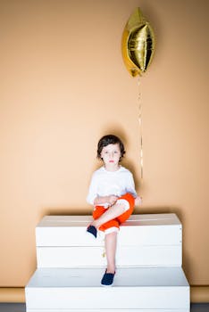 孩子戴着金色气球，坐在白色台阶上，背景是中性的