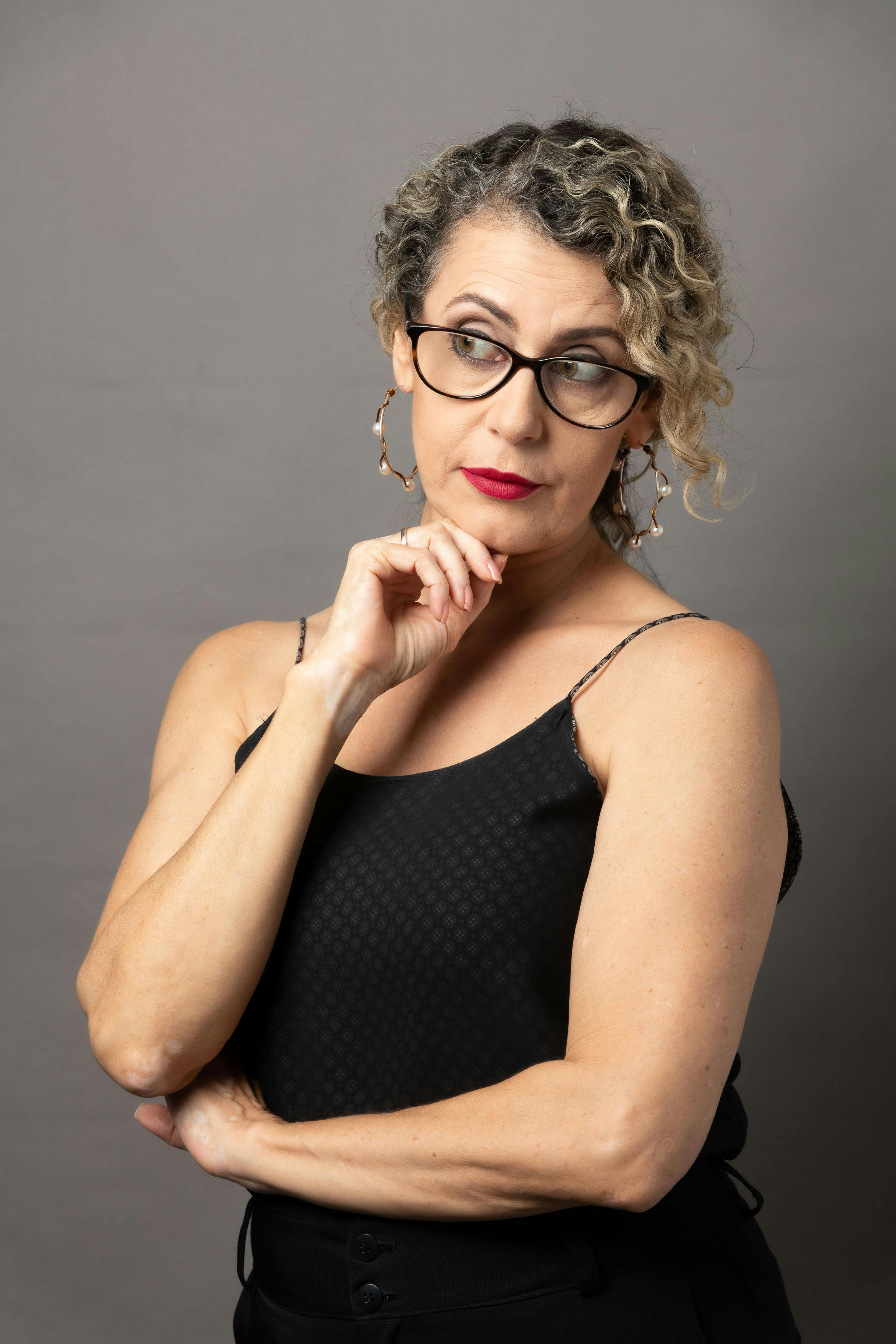 Portrait of a thoughtful woman with curly hair and glasses against a gray background.