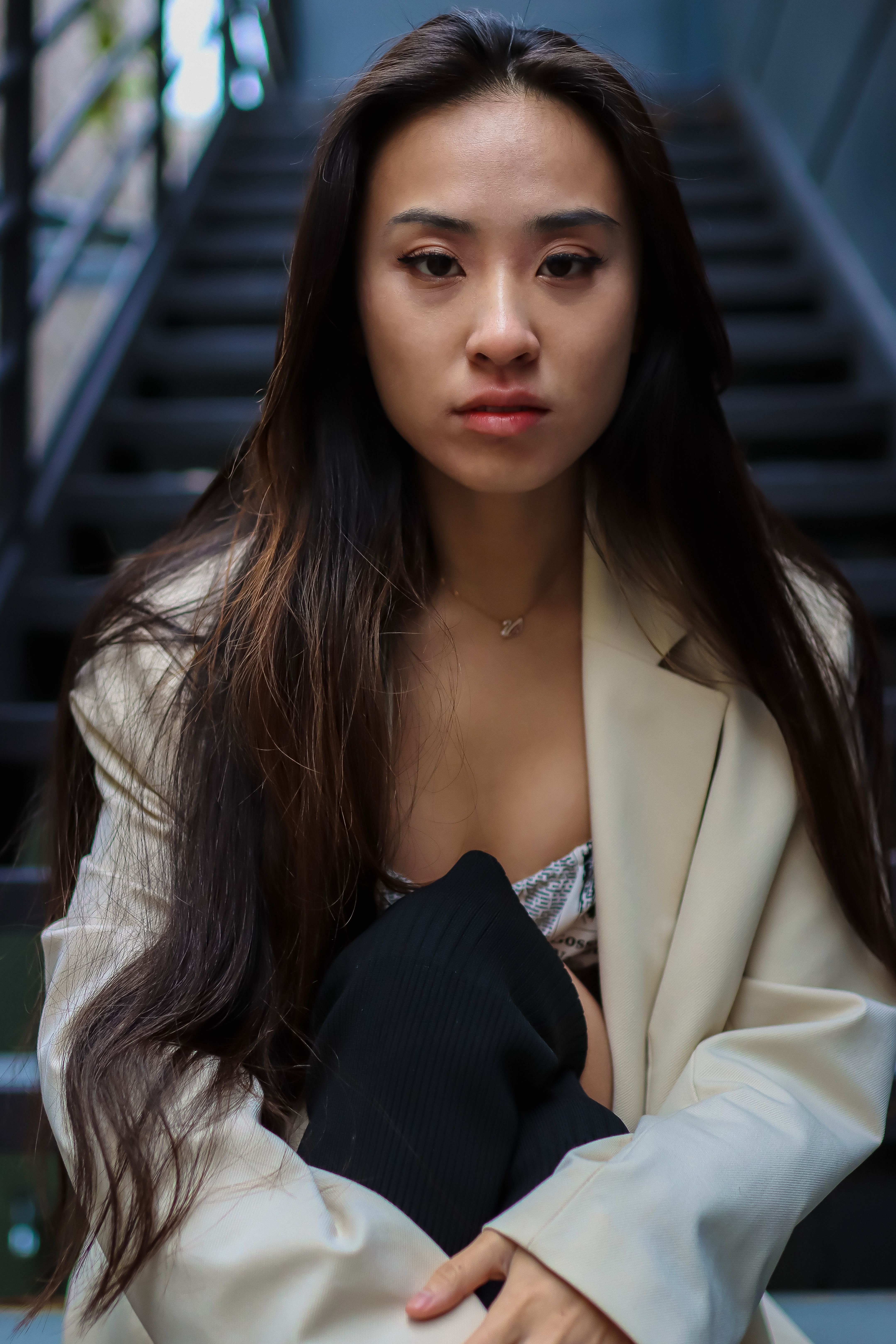 Thoughtful woman in blazer seated on stairs, urban backdrop.