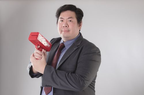 Free Confident businessman posing with a red money gun in a studio setting, wearing a formal suit. Stock Photo