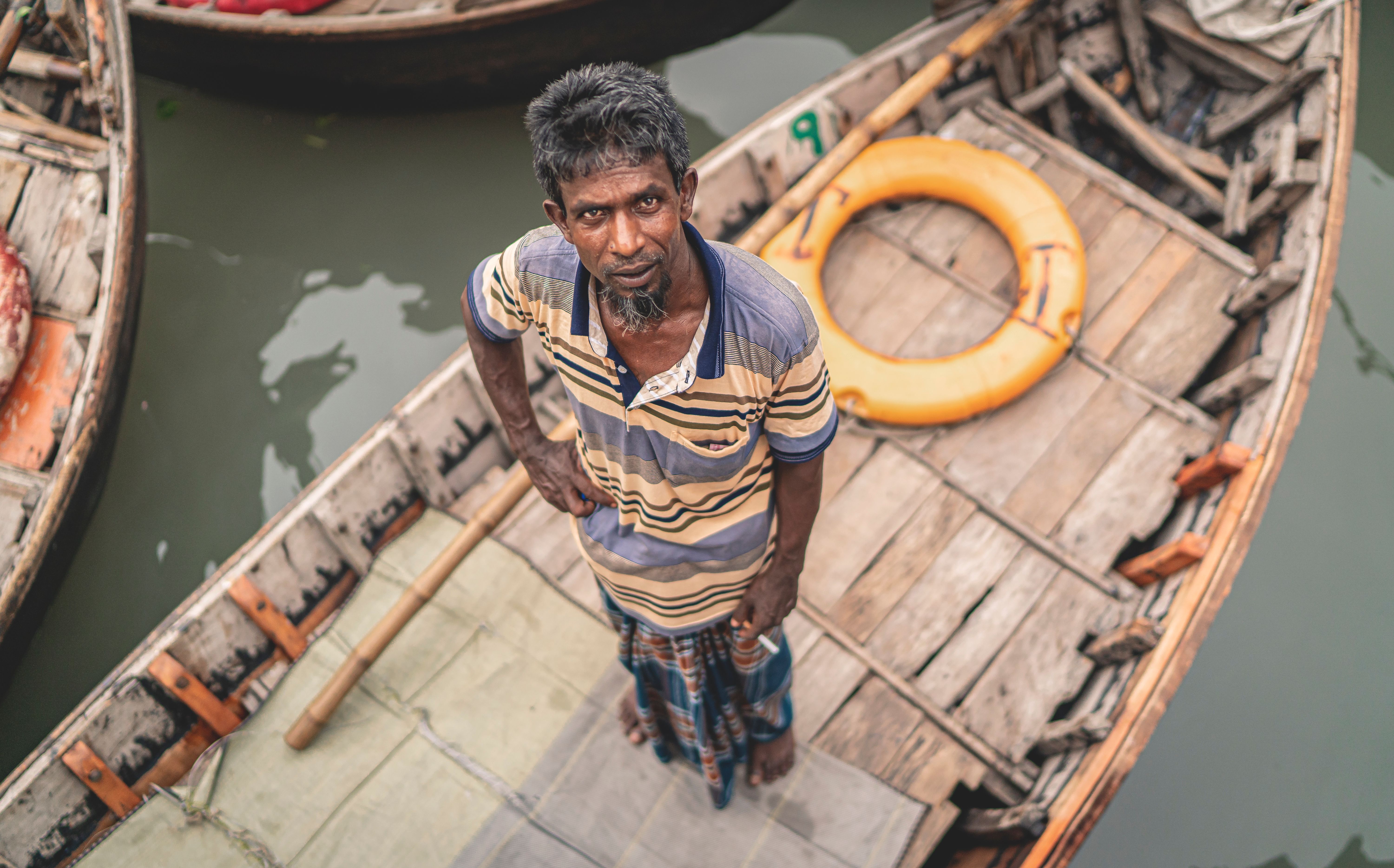 A man standing on a wooden boat in Dhaka, Bangladesh, looking up at the camera.