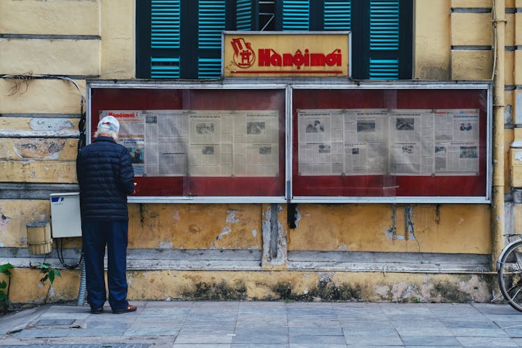 Elderly Man Reading A Newspaper In A Display Case On The Wall Of An Old Building